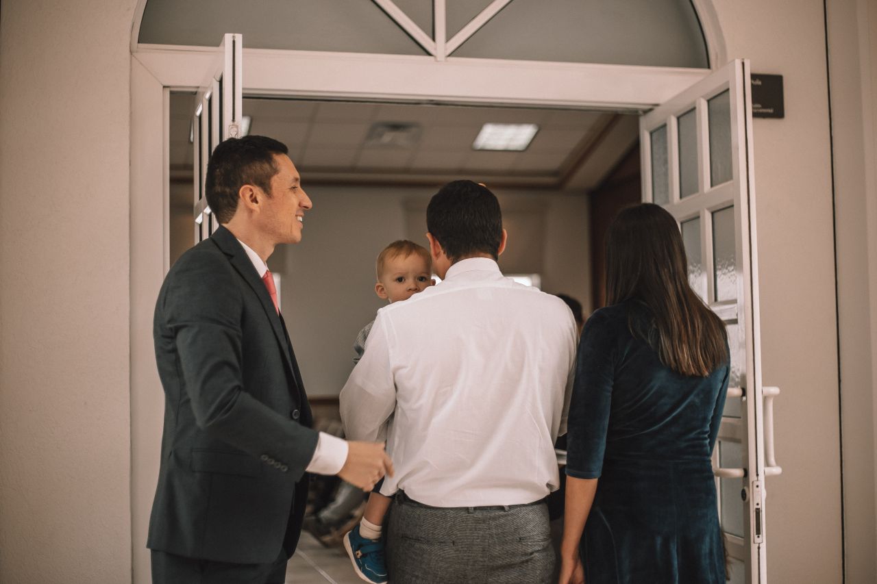 Um homem segura uma porta aberta para uma jovem família entrar em um edifício da Igreja para aprender sobre o evangelho de Jesus Cristo.